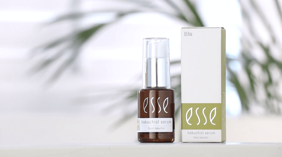 A bottle of Esse Bakuchiol Serum with a clear cap stands beside its matching box, both labeled "Esse Bakuchiol Serum," set against a softly blurred background featuring hints of green leaves.