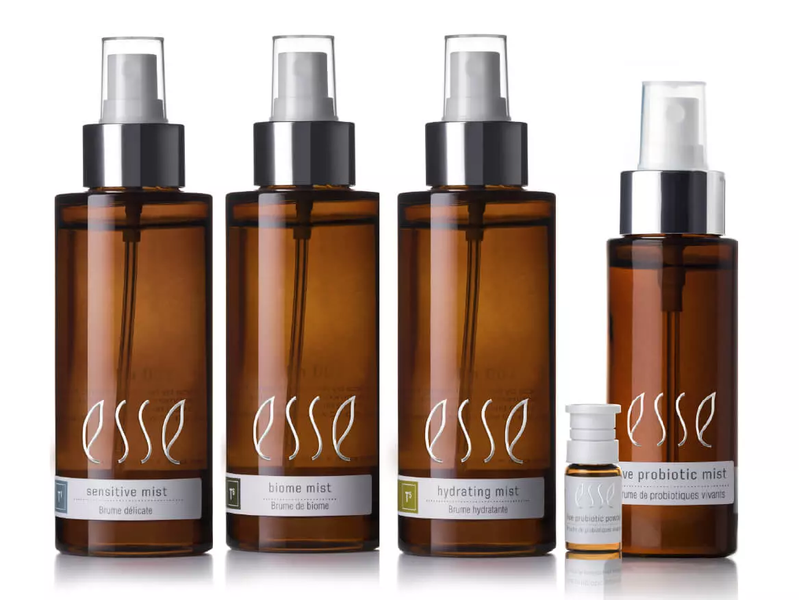 Four amber spray bottles labeled "esse" stand in a row, each with a unique mist—sensitive, biome, hydrating, and live probiotic—to support het microbioom na esthetische behandeling. A small vial sits in front of the last bottle.