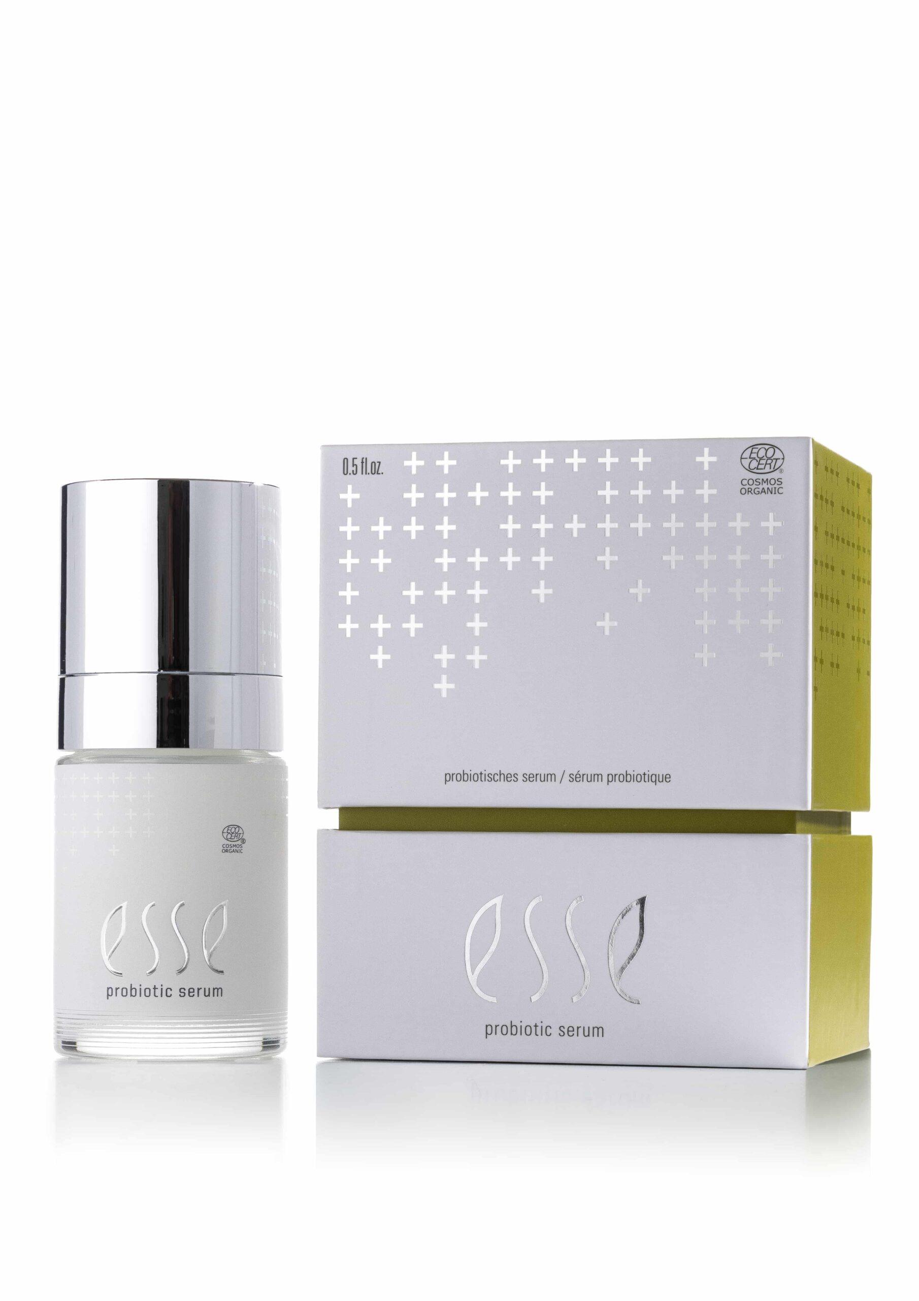 A bottle of Esse Probiotic Serum, topped with a shiny silver cap, stands beside its white and yellow box adorned with silver cross symbols and product details.