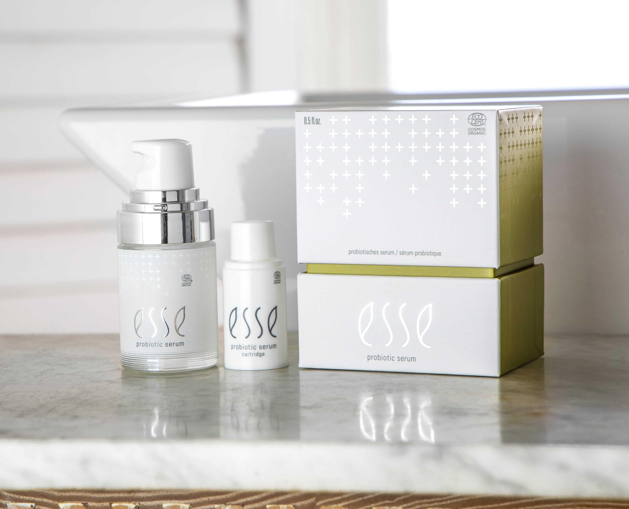 On a marble countertop by a window, an Esse Probiotic Serum glass pump bottle, a small plastic bottle, and two stacked serum boxes with white packaging and silver-green accents are displayed.
