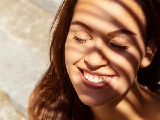 A young woman with brown hair smiles with her eyes closed as sunlight filters through window blinds, casting striped shadows across her face—a moment of pure bliss enhanced by levende probiotische huidverzorging.
