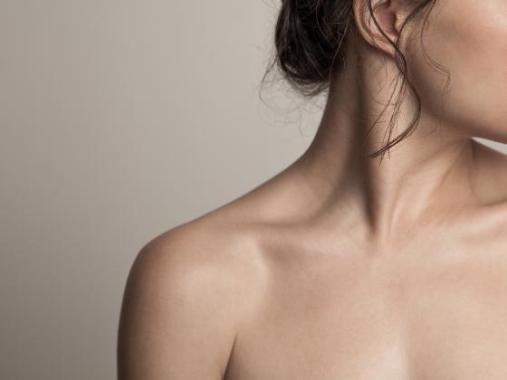 Close-up of a woman's bare shoulder, neck, and lower face, with wisps of brown hair and soft lighting against a neutral background—capturing the natural glow that comes from de vitamine D die je niet uit een pil haalt.